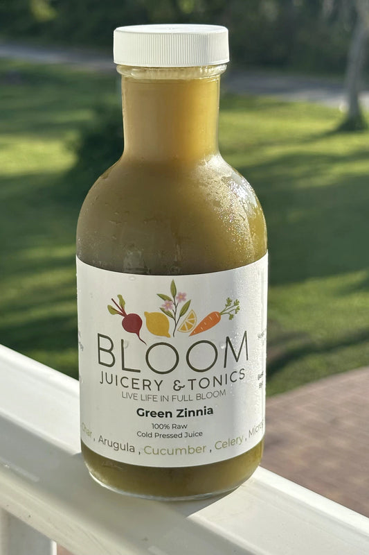 Bottle of Bloom Juicery & Tonics Green Zinnia juice on a windowsill with a garden background.